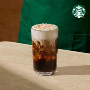 A tall glass filled with iced coffee, topped with whipped cream and sprinkled with toasted coconut flakes, sits on a light-colored table. A green apron is partially visible in the background.