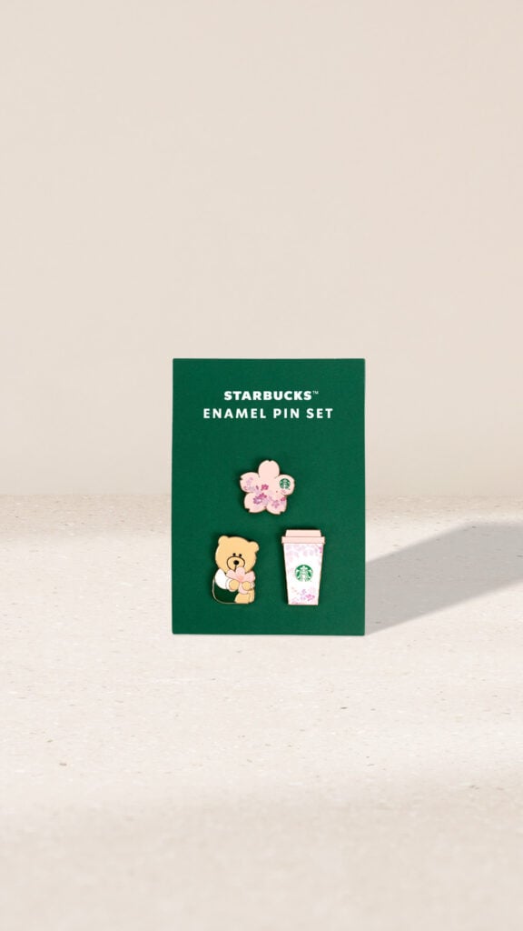 A green card displays a Starbucks enamel pin set featuring three pins: a bear holding flowers, a pink flower, and a pink Starbucks cup.