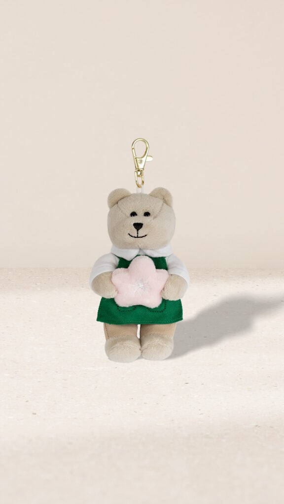 A cute plush bear keychain wearing a green apron and white shirt, holding a pink star-shaped object. The background is neutral.