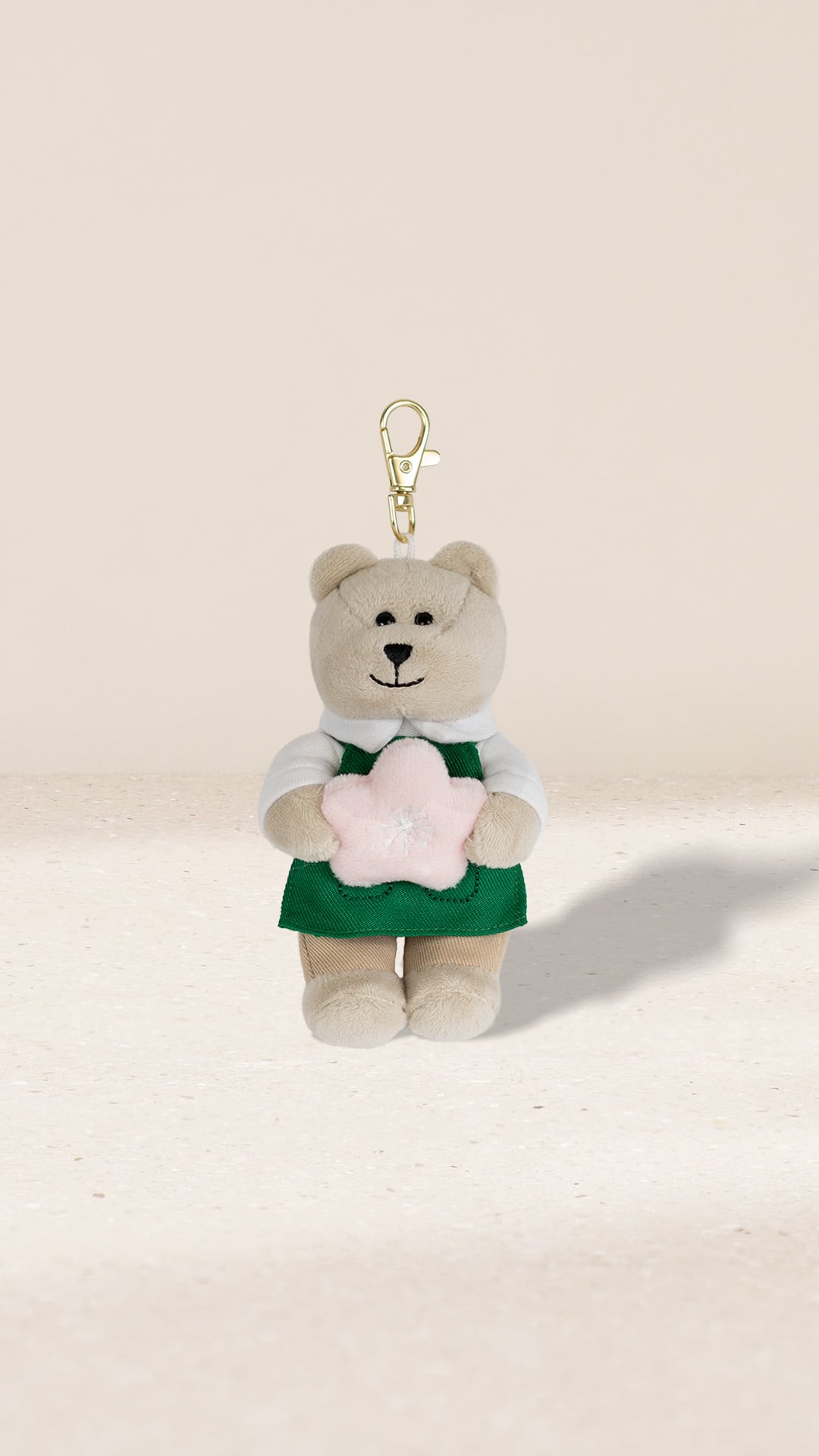 A cute plush bear keychain wearing a green apron and white shirt, holding a pink star-shaped object. The background is neutral.