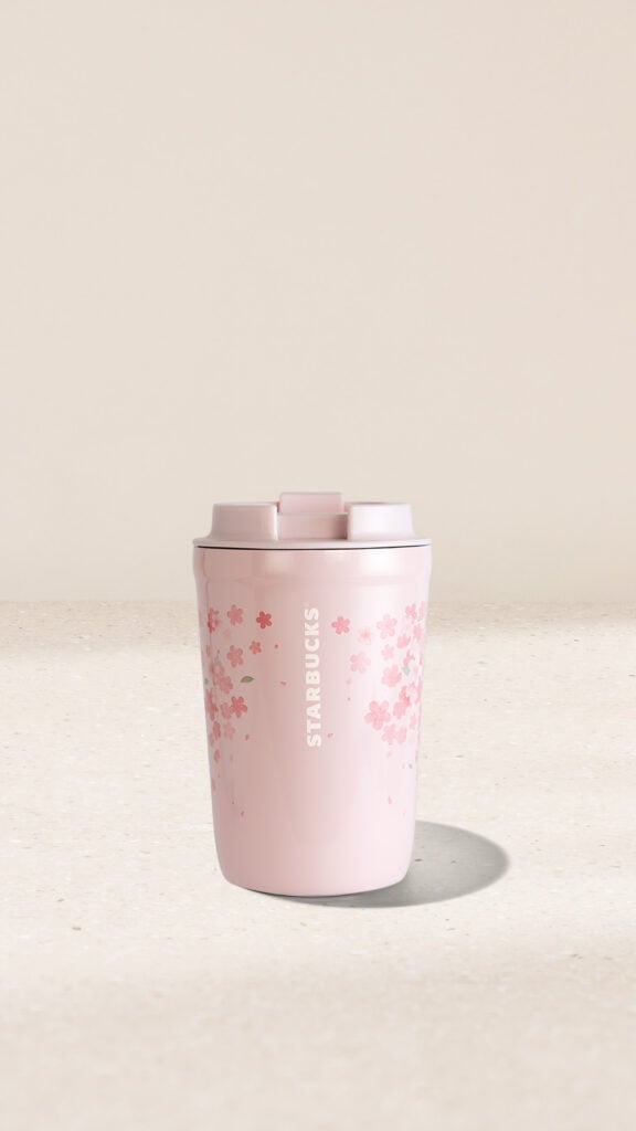 A pink Starbucks travel mug adorned with cherry blossom designs, featuring a secure lid and a smooth finish, sits on a light surface.