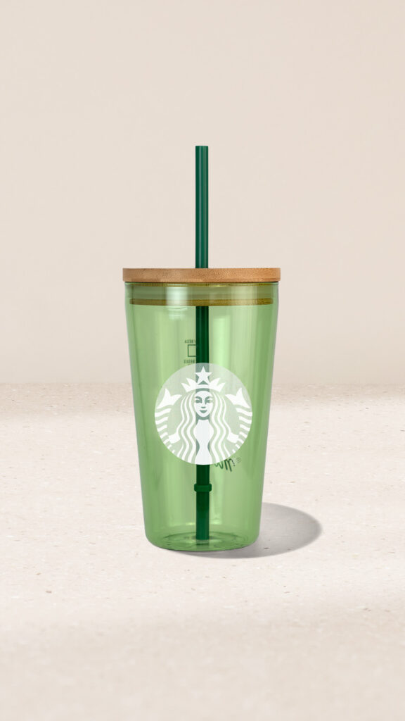 A green reusable cup with a bamboo lid and a green straw, featuring a white logo on the front. The background is neutral.