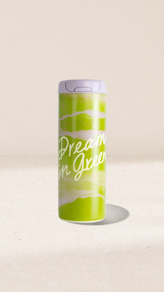 A green travel mug with a purple lid features the phrase "Dream in Green" in white lettering, set against a light background.