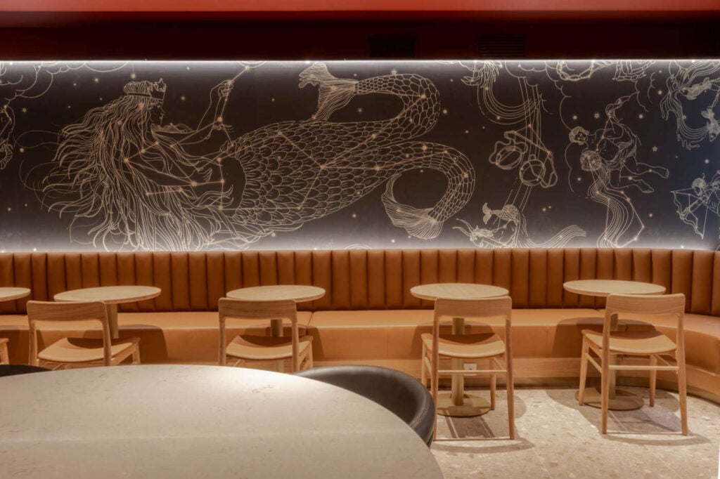 A cozy café interior features a mural of a mermaid and celestial designs on a dark wall. Lighted seating and wooden tables enhance the ambiance.