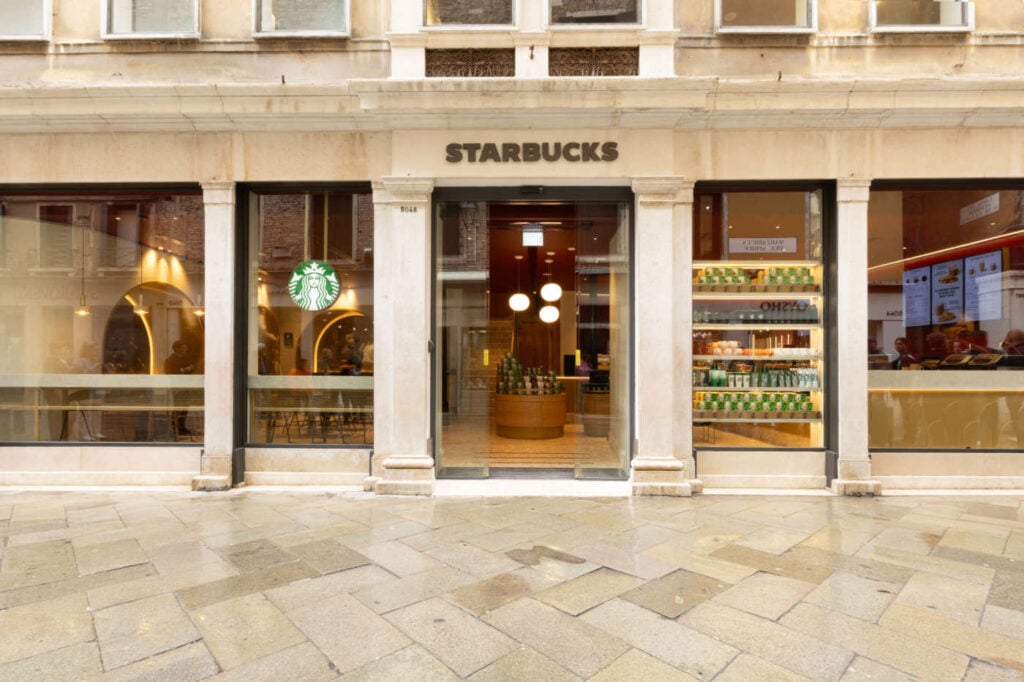 A Starbucks storefront featuring large glass windows, a prominent logo, and a display of products inside. The entrance is framed by light-colored stone.