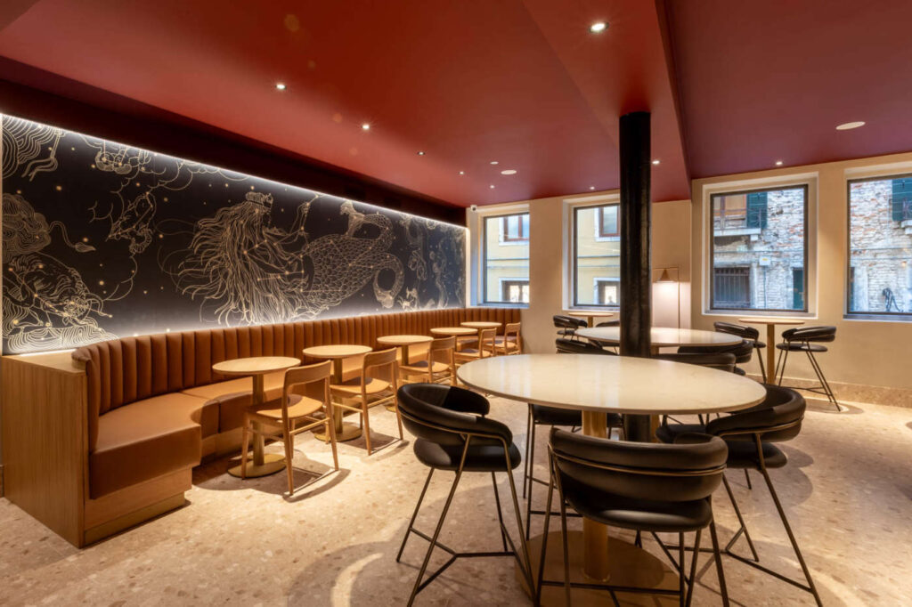 A modern café interior featuring a curved brown banquette, round wooden tables, and black chairs. A large mural with intricate designs adorns the wall, illuminated by soft lighting. Large windows provide natural light, enhancing the inviting atmosphere.