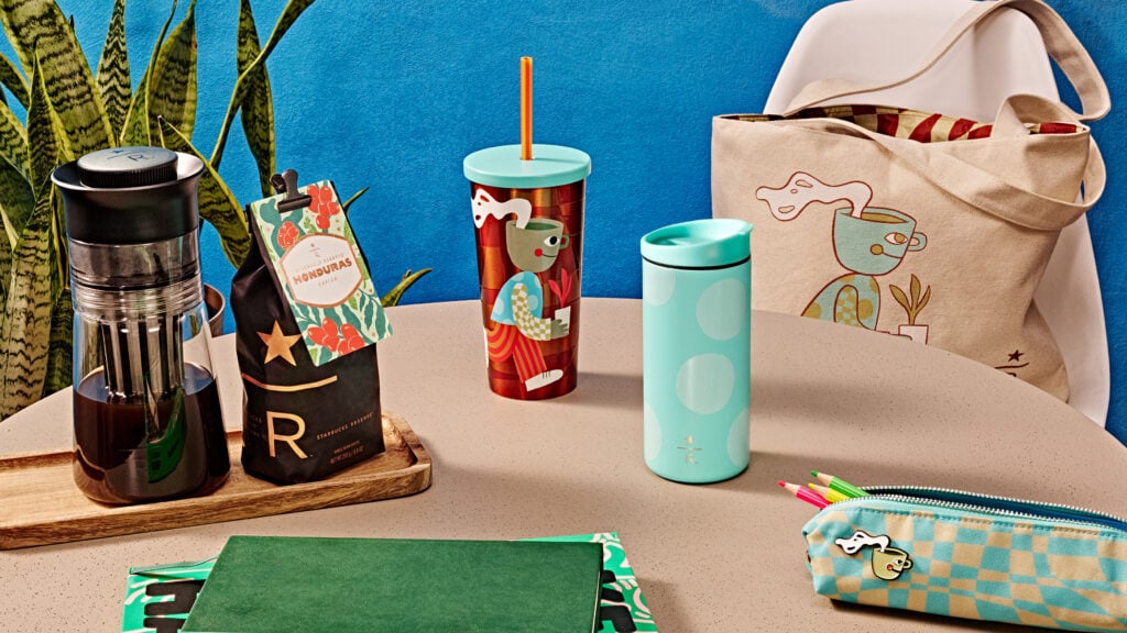 A colorful table setting features a tall cup with a playful character design and an orange straw, alongside a mint green travel mug with polka dots. A bag of coffee and a green notebook are also visible, adding to the vibrant atmosphere.