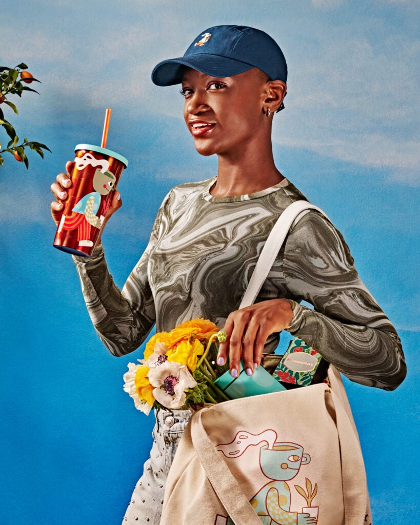 A person holds a colorful drink cup and a tote bag filled with flowers and items. They wear a patterned long-sleeve shirt and a cap, with a blue background.