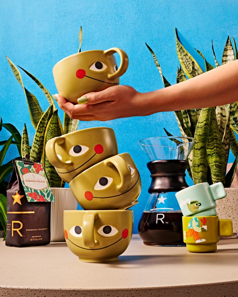 A hand is stacking playful, smiling green mugs on a light surface, with a coffee maker and coffee bag nearby, surrounded by plants.