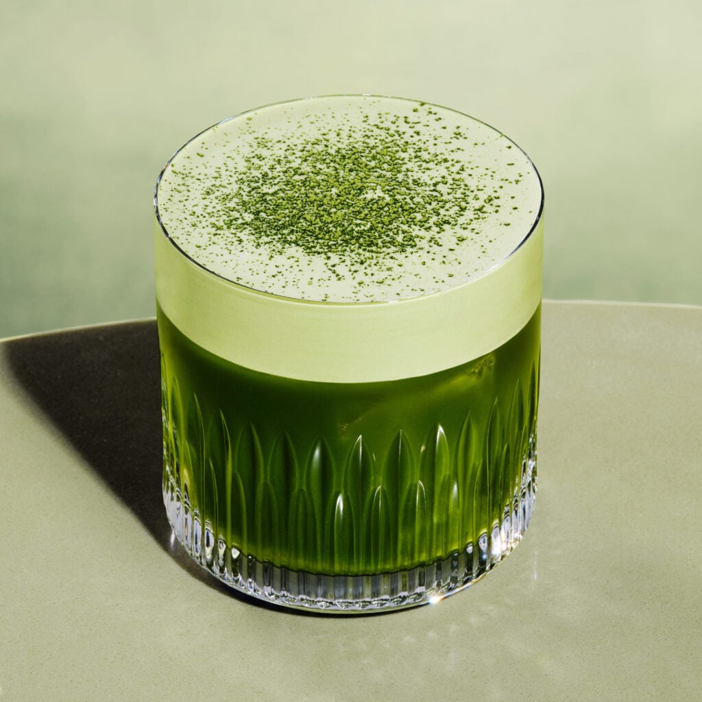 A glass filled with a vibrant green drink, topped with a frothy layer and sprinkled with green powder, sits on a smooth surface.