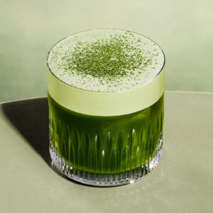 A glass filled with a vibrant green drink, topped with a frothy layer and sprinkled with green powder, sits on a smooth surface.