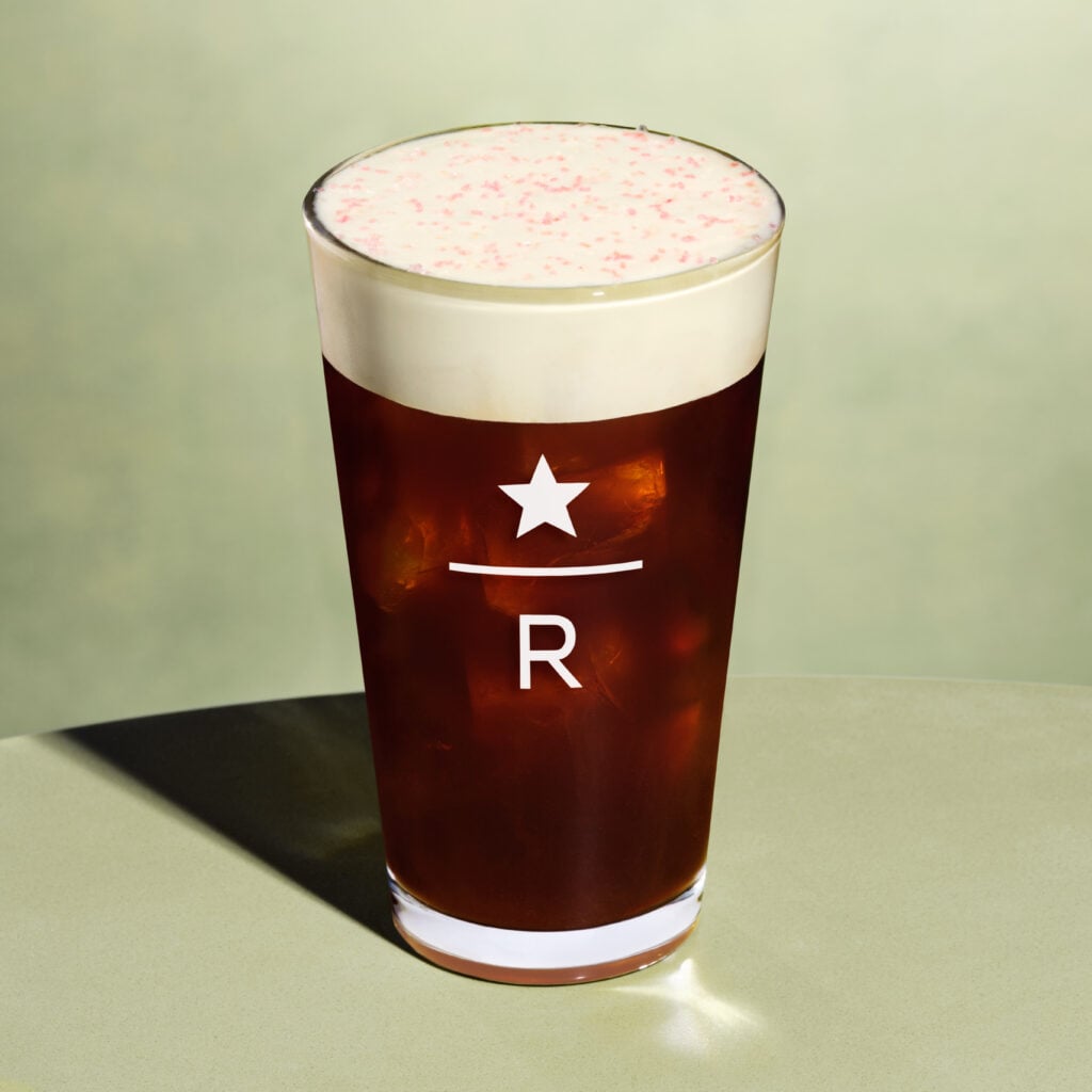 A clear glass filled with iced coffee topped with a creamy foam and pink sprinkles. The glass features a logo with a star and the letter "R.