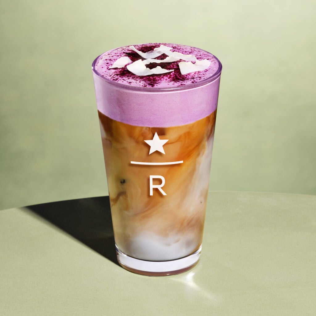 A tall glass features a layered drink with a creamy purple foam on top, sprinkled with coconut flakes, and a mix of brown and white liquid below.