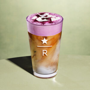 A tall glass features a layered drink with a creamy purple foam on top, sprinkled with coconut flakes, and a mix of brown and white liquid below.