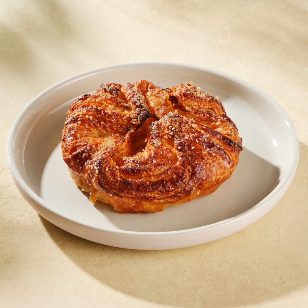 A golden-brown pastry with a flaky, spiral shape sits on a round, light-colored plate. The surface is slightly shiny and sprinkled with coarse sugar.