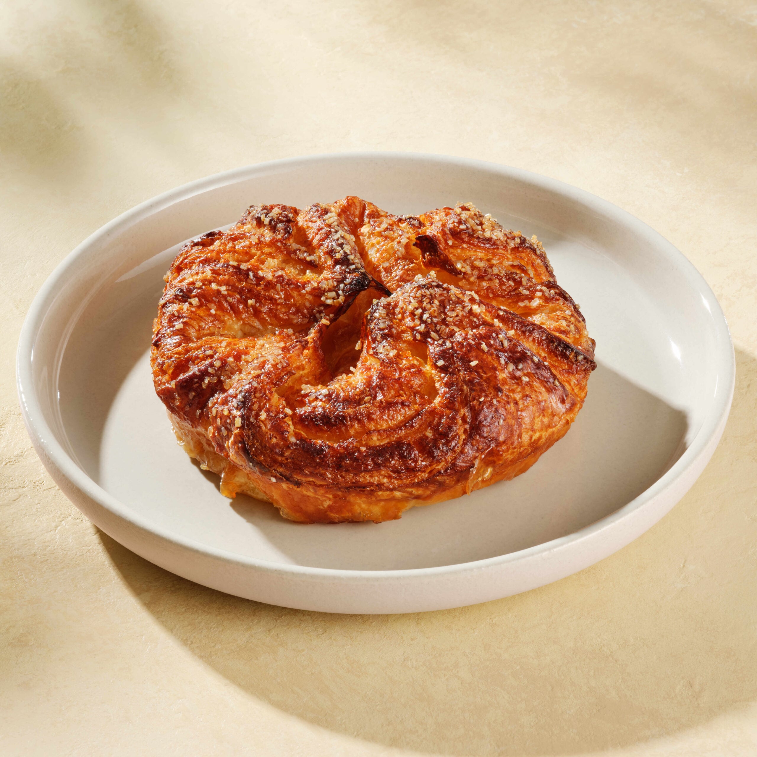 A golden-brown pastry with a flaky, spiral shape sits on a round, light-colored plate. The surface is slightly shiny and sprinkled with coarse sugar.