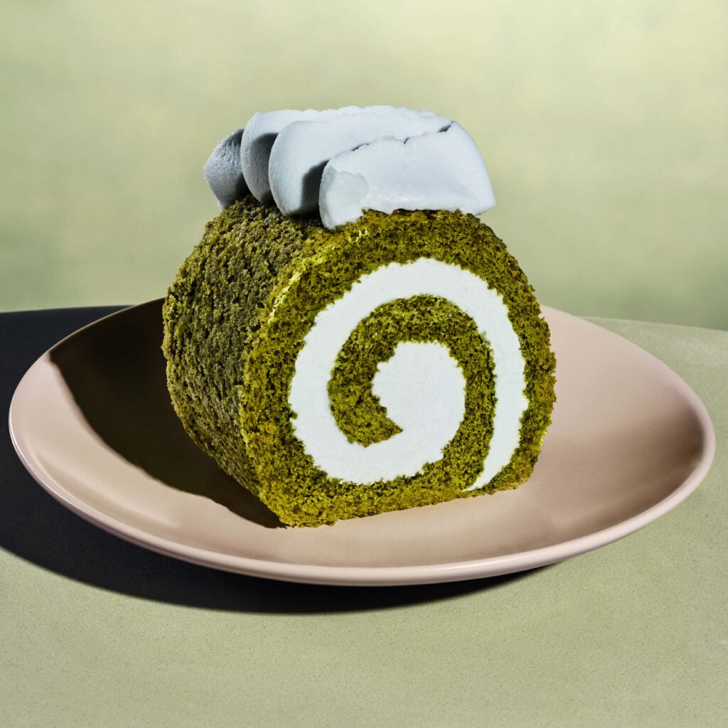 A slice of green matcha roll cake is displayed on a pale pink plate. The cake features a white cream filling and is topped with a dollop of whipped cream.