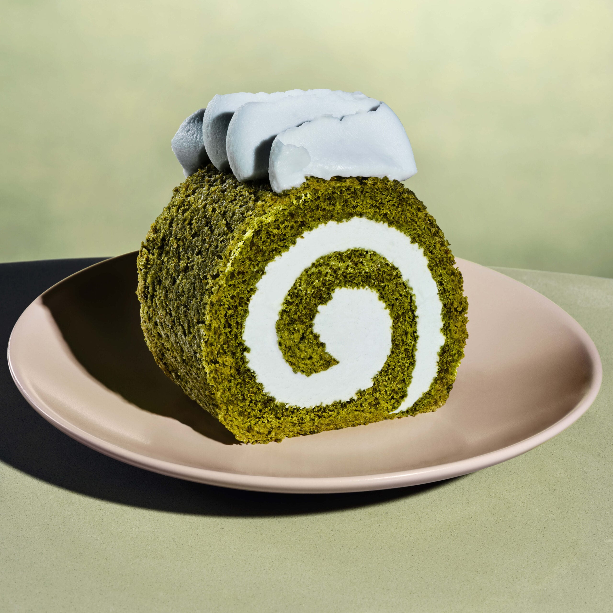 A slice of green matcha roll cake is displayed on a pale pink plate. The cake features a white cream filling and is topped with a dollop of whipped cream.