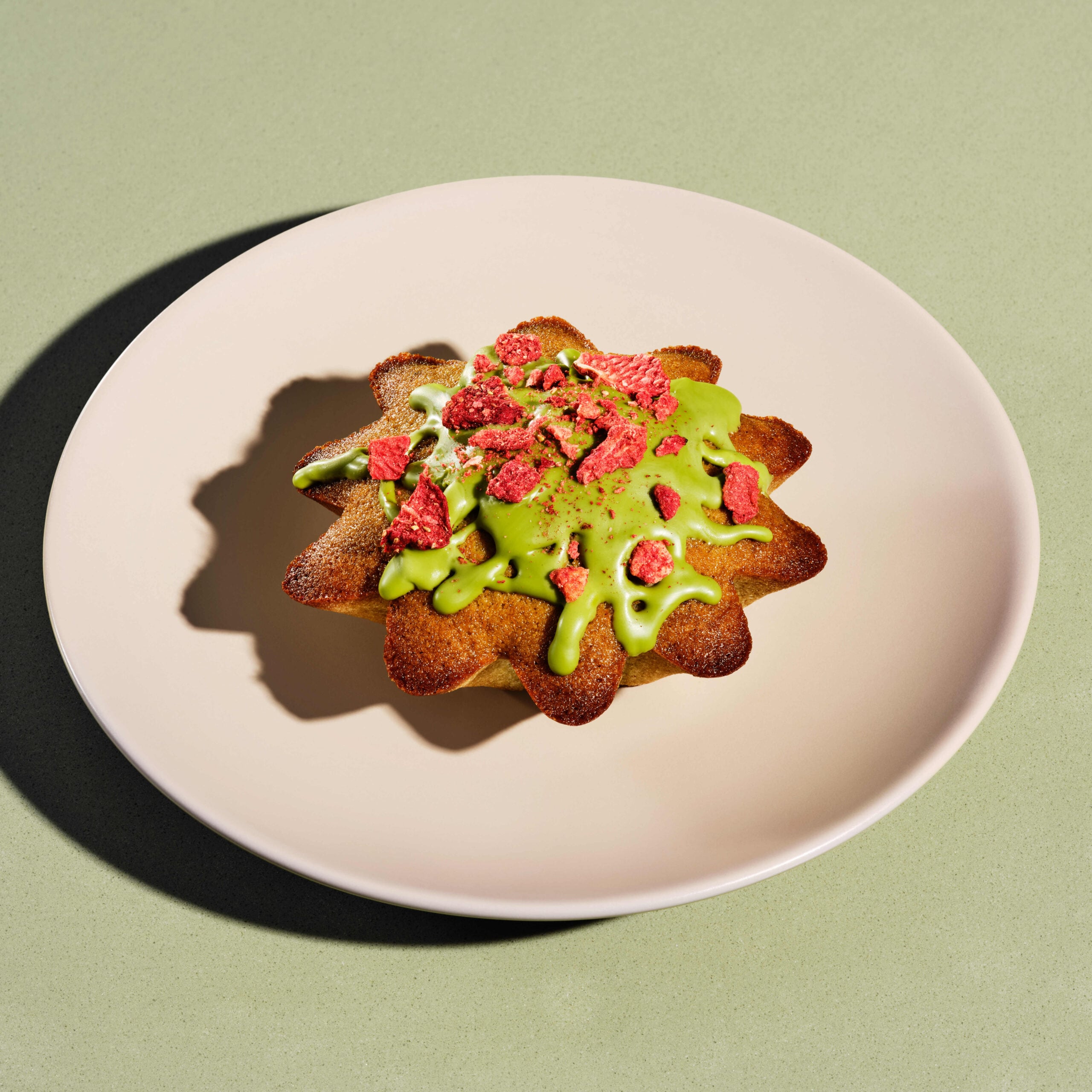A star-shaped pastry topped with green icing and crushed red strawberries, placed on a pale plate against a green background.