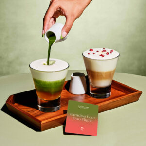 A hand pours green liquid into a layered drink with green, orange, and white hues. Next to it is a creamy drink topped with pomegranate seeds, on a wooden tray with a card labeled "Paradise Pour Duo Flight.