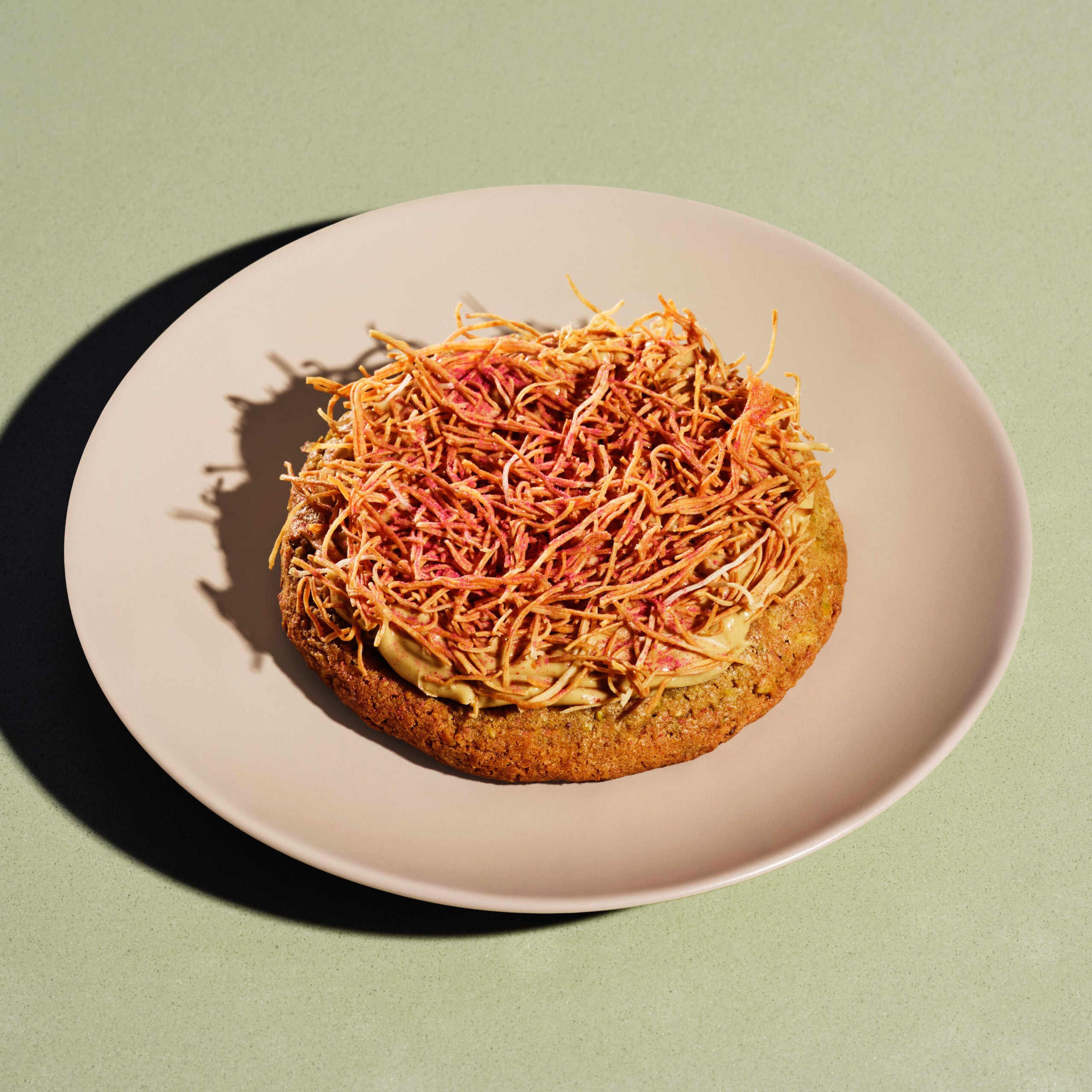A round, thick cookie topped with a layer of light brown frosting and finely shredded, crispy strands, served on a pale pink plate.