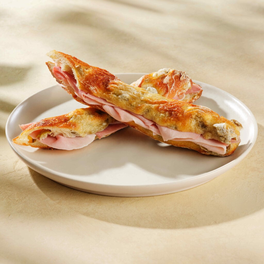 Two toasted sandwich rolls filled with slices of ham are arranged on a round, white plate. The rolls have a golden-brown crust.