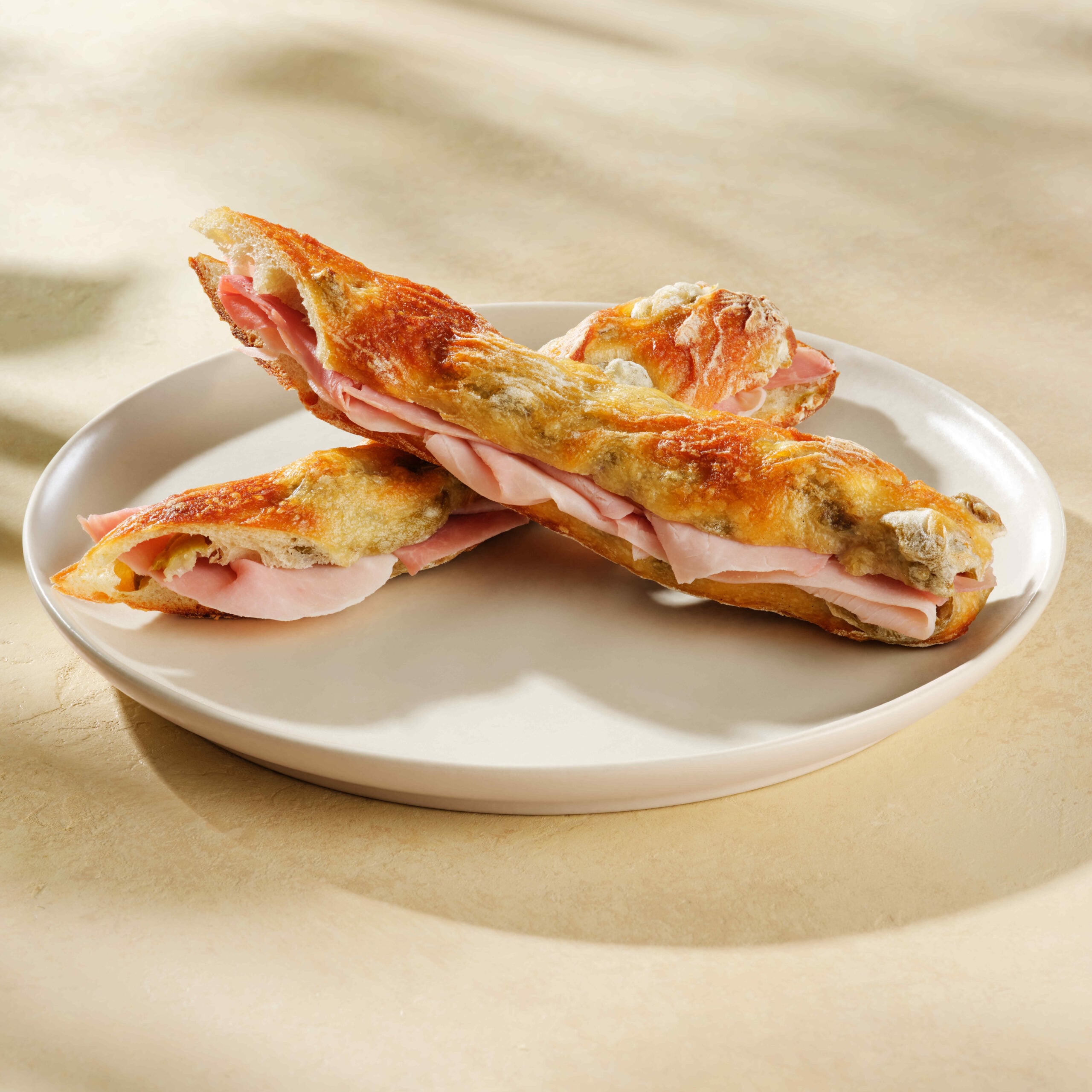 Two toasted sandwich rolls filled with slices of ham are arranged on a round, white plate. The rolls have a golden-brown crust.
