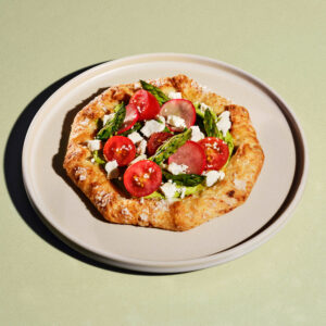 A rustic, octagonal tart features a golden-brown crust topped with green spread, cherry tomatoes, asparagus, radish slices, and crumbled feta cheese. It's served on a light-colored plate against a soft green background.