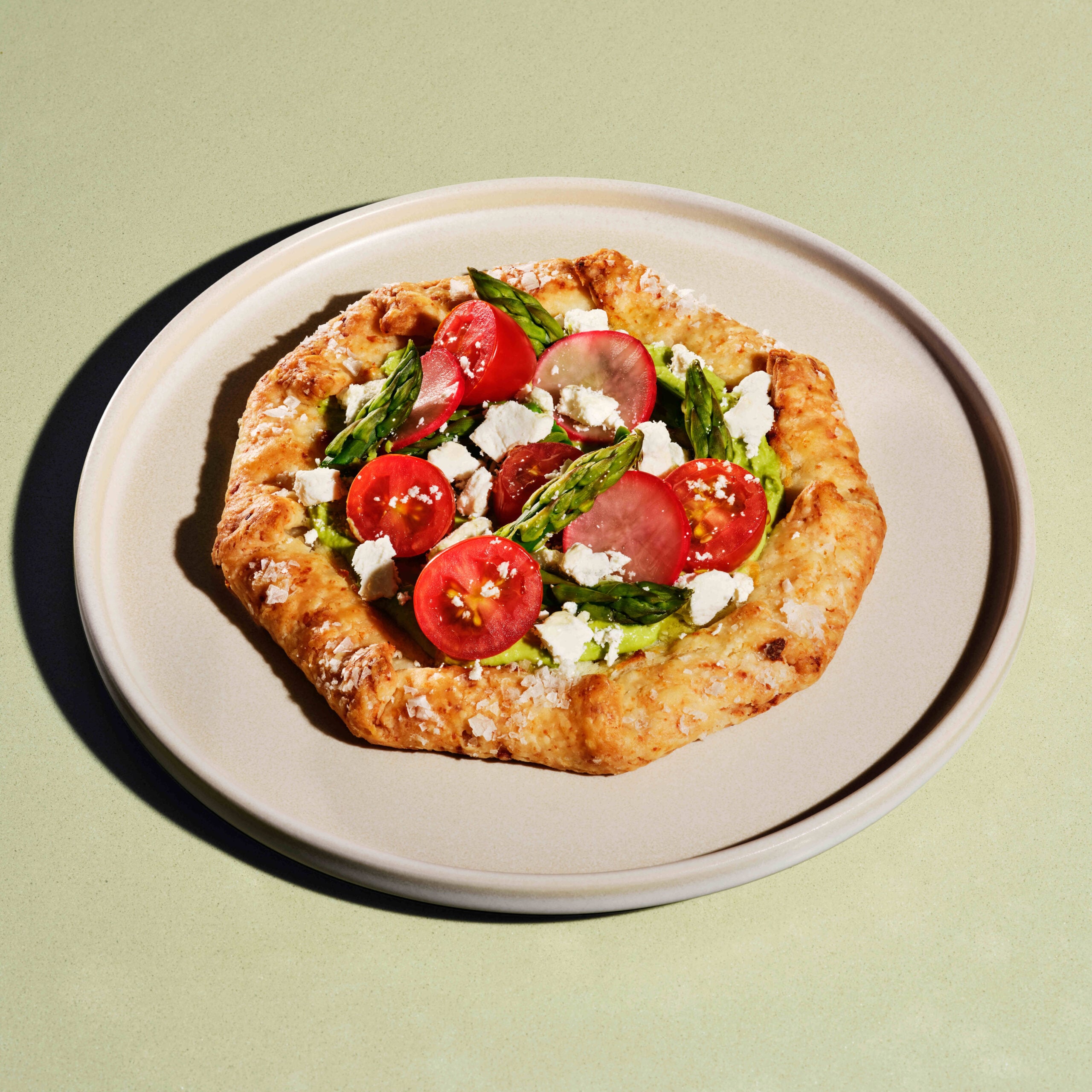 A rustic, octagonal tart features a golden-brown crust topped with green spread, cherry tomatoes, asparagus, radish slices, and crumbled feta cheese. It's served on a light-colored plate against a soft green background.