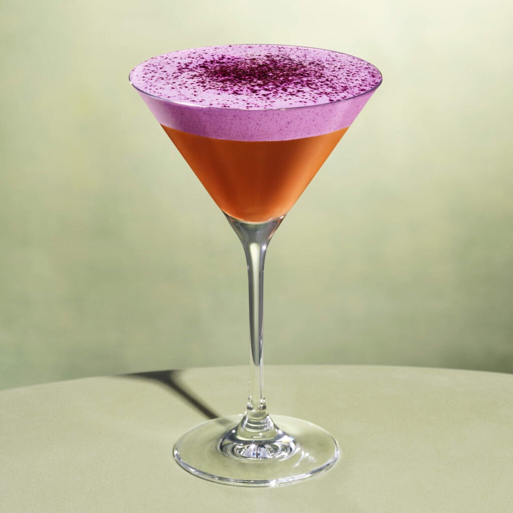 A stylish cocktail in a martini glass features a vibrant orange base topped with a frothy purple layer and sprinkled with dark powder.