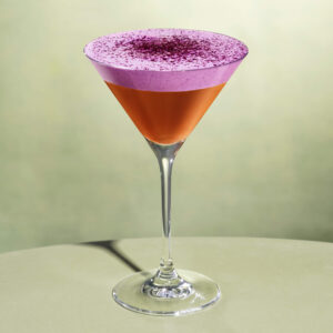 A stylish cocktail in a martini glass features a vibrant orange base topped with a frothy purple layer and sprinkled with dark powder.