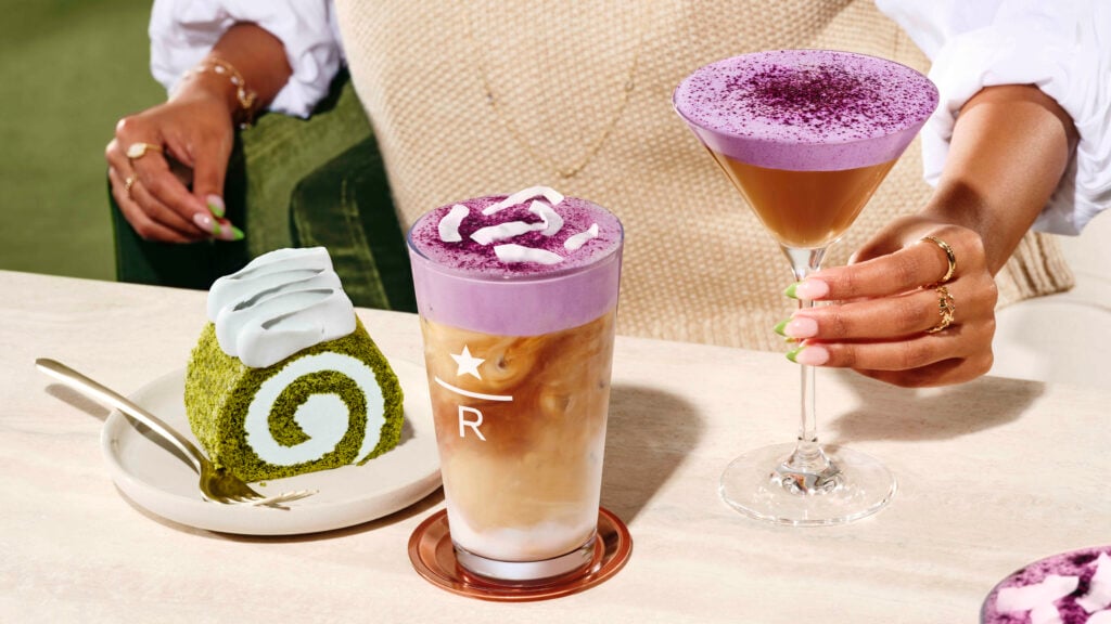 A vibrant purple drink topped with coconut flakes sits next to a green roll cake on a plate. A second purple drink in a martini glass is held by a hand.