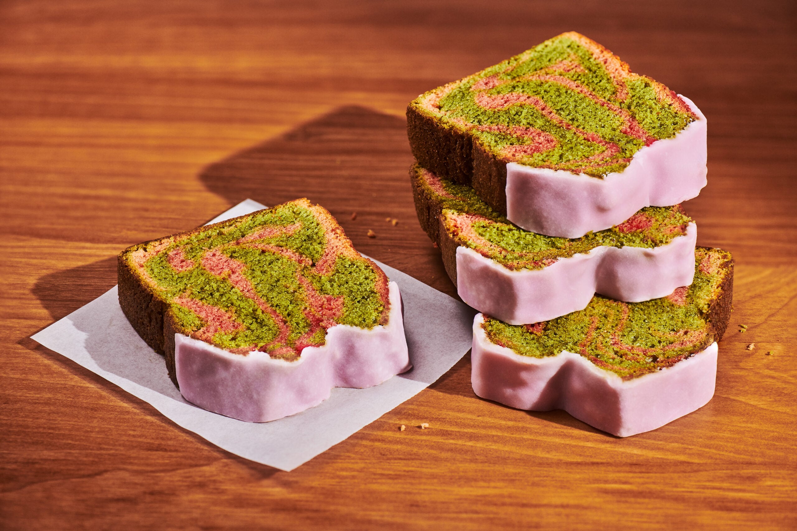 Colorful slices of cake with green and pink swirls, topped with a pink icing, are stacked on a wooden surface.