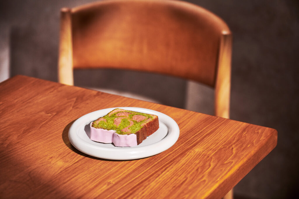 A slice of colorful cake with green and pink swirls sits on a white plate, placed on a wooden table. A wooden chair is visible in the background.