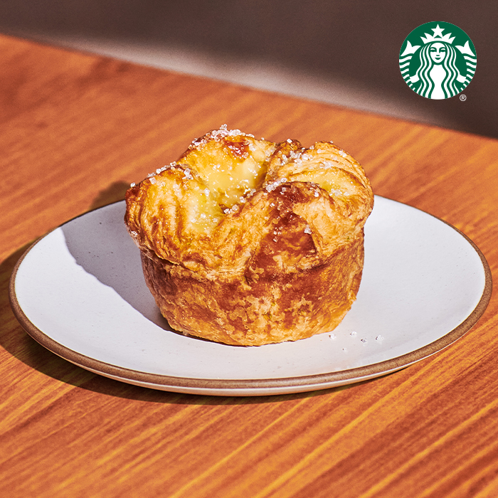 A golden-brown pastry sits on a white plate with a thin rim. The pastry has a flaky texture and a creamy filling, topped with a sprinkle of sugar. The background features a wooden surface.