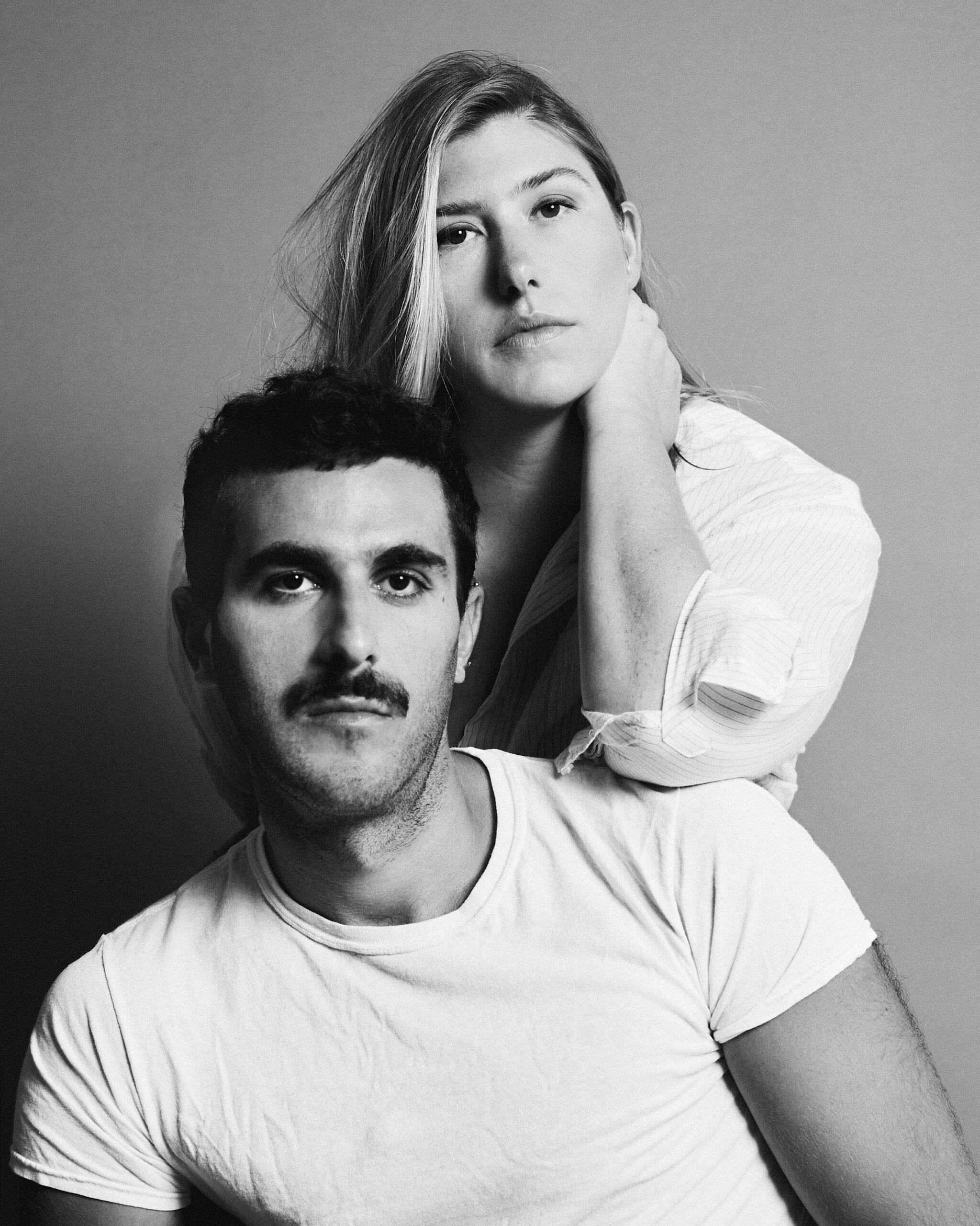 A black-and-white portrait of a man and a woman. The man, with a mustache, sits in front, while the woman stands behind him, resting her hand on his shoulder. Both have serious expressions, and the background is a soft gray.