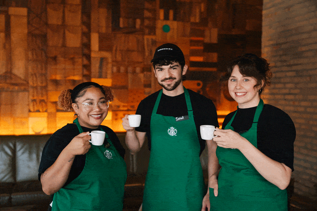 Three baristas in green aprons hold small cups, smiling at the camera. The background features warm lighting and textured walls.