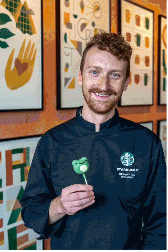 A smiling man in a black chef's jacket holds a green frog-shaped lollipop. Colorful abstract art is displayed on the wall behind him.