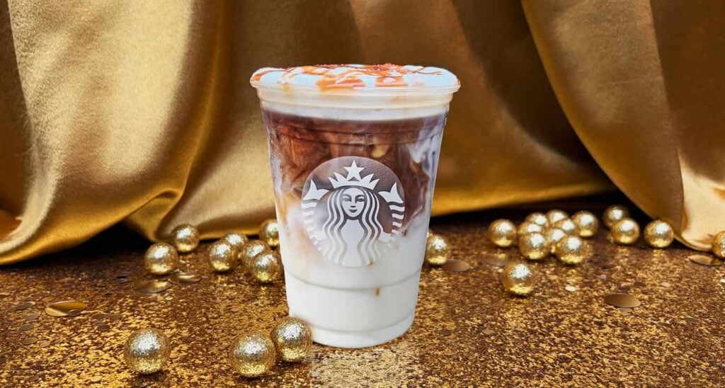A clear cup filled with a layered coffee drink featuring milk and dark coffee, topped with caramel drizzle. It's set against a golden backdrop with shiny gold balls scattered around.