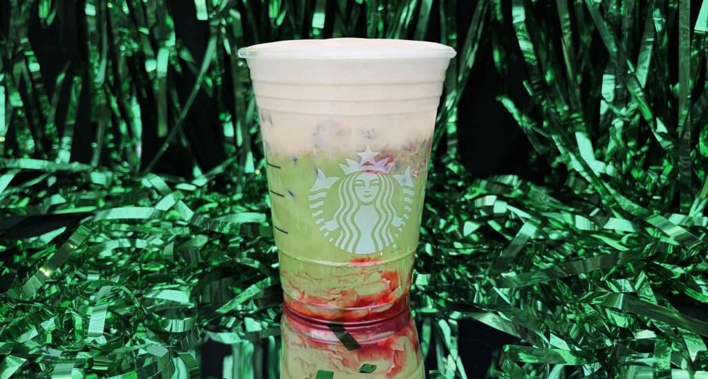 A Starbucks cup filled with a layered drink featuring green and pink hues, set against a backdrop of shiny green decorations.
