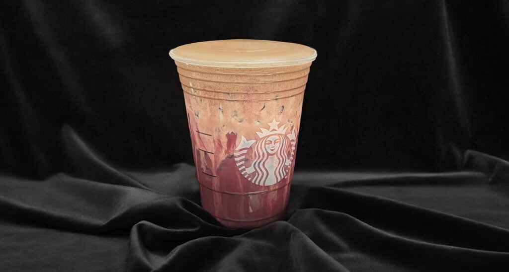 A clear cup filled with a layered iced beverage, featuring a creamy top and colorful swirls of liquid, set against a dark fabric background.
