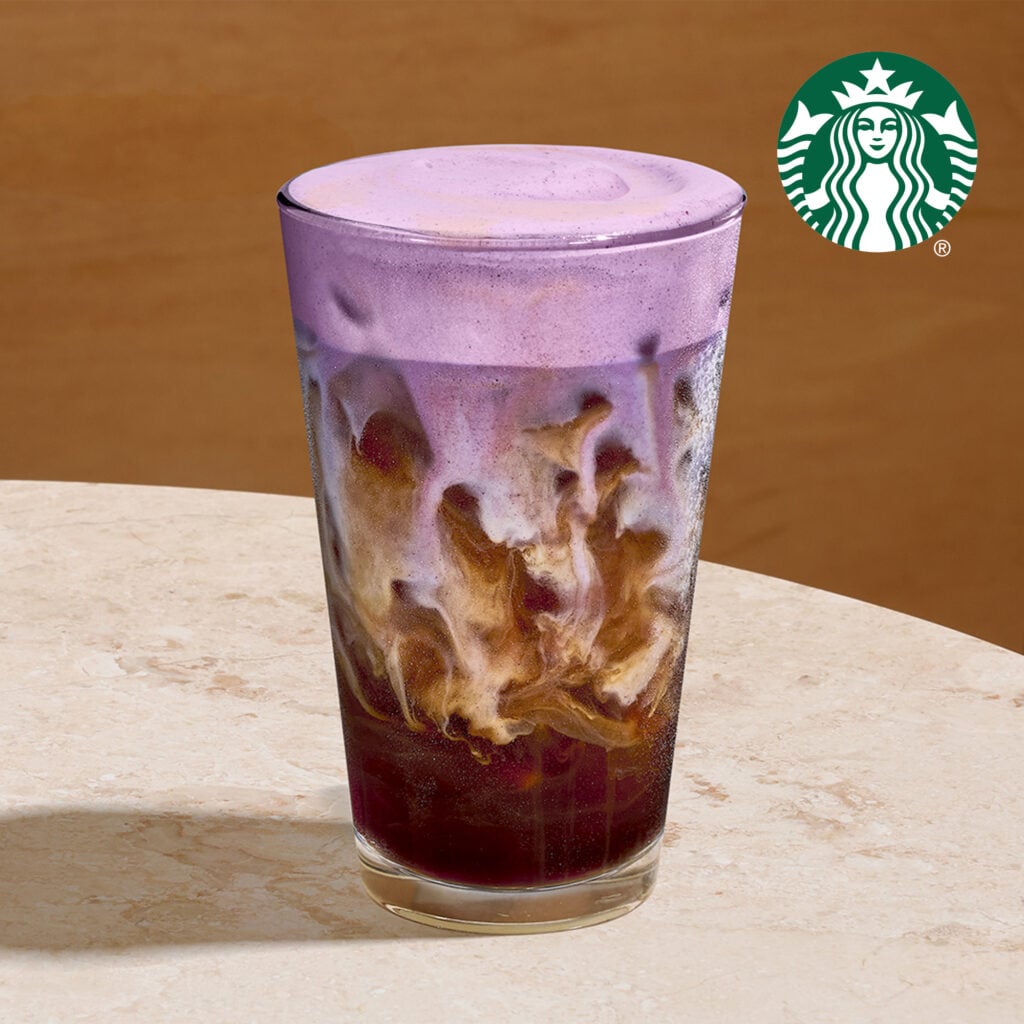 A tall glass filled with a layered drink featuring a purple frothy top, swirling with dark coffee and cream below.