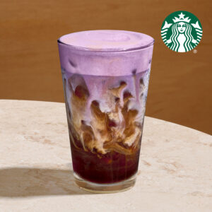 A tall glass filled with a layered drink featuring a purple frothy top, swirling with dark coffee and cream below.