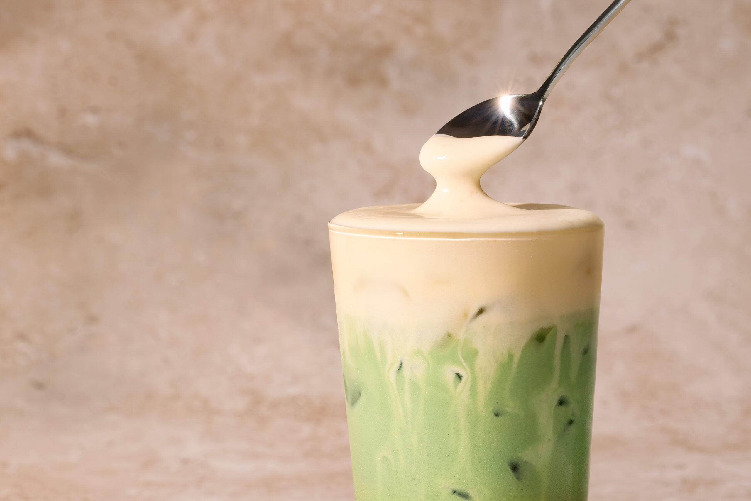 A tall glass filled with layered green matcha and creamy beige liquid, topped with a glossy foam. A spoon is poised above, dripping.