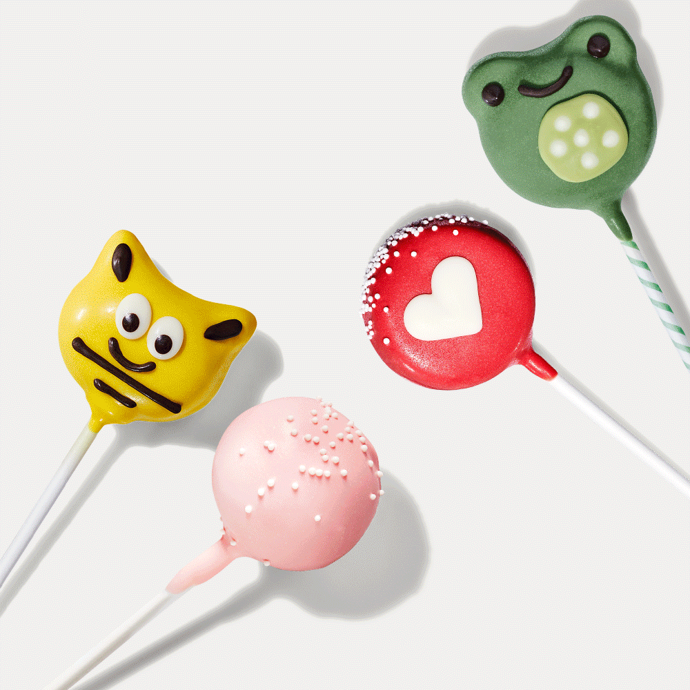 Four colorful cake pops are displayed on white sticks. They include a yellow cat face, a green frog, a pink ball with sprinkles, and a red heart-shaped pop.