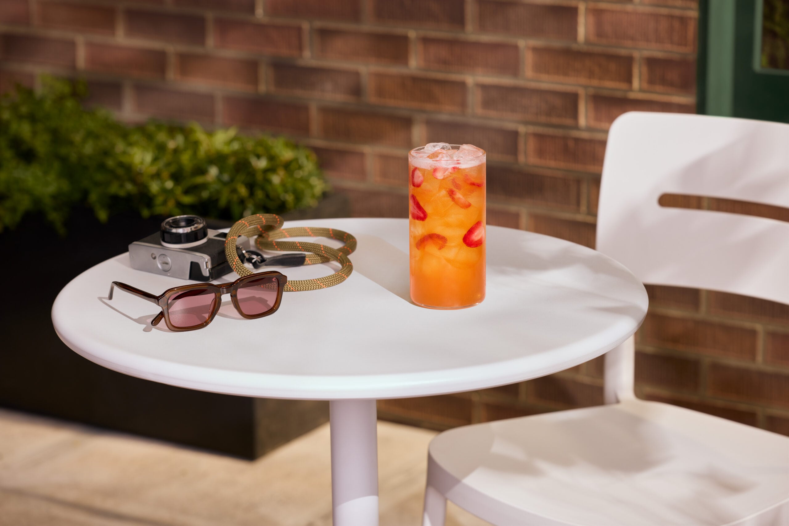 A tall glass filled with a colorful drink featuring ice and strawberry slices sits on a white table. Nearby are brown sunglasses and a vintage camera. A green plant is visible in the background against a brick wall.
