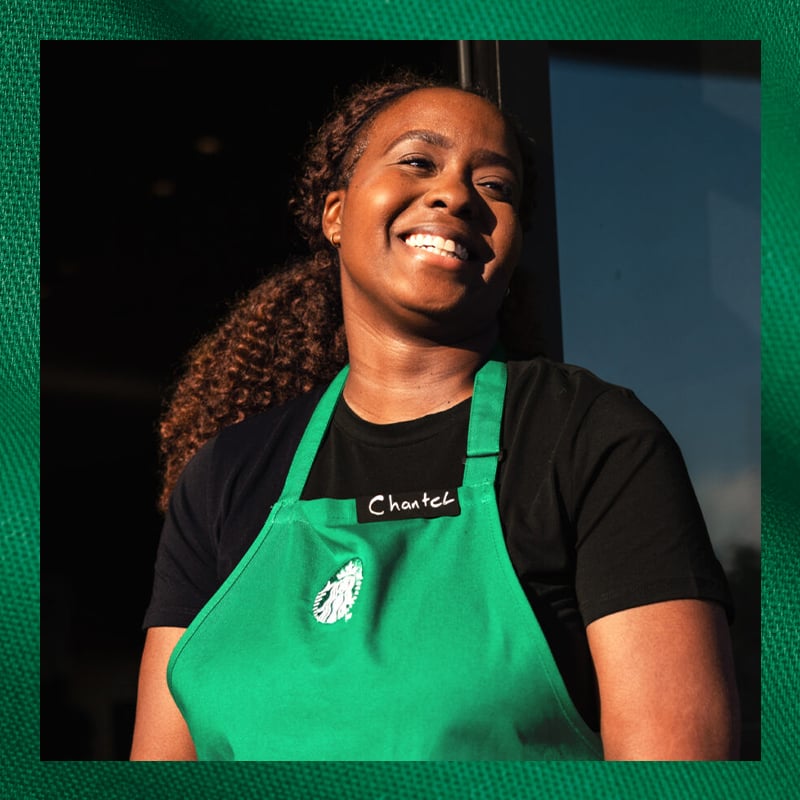 A smiling woman with curly hair wears a green apron over a black shirt. She stands in front of a window, exuding a friendly demeanor.