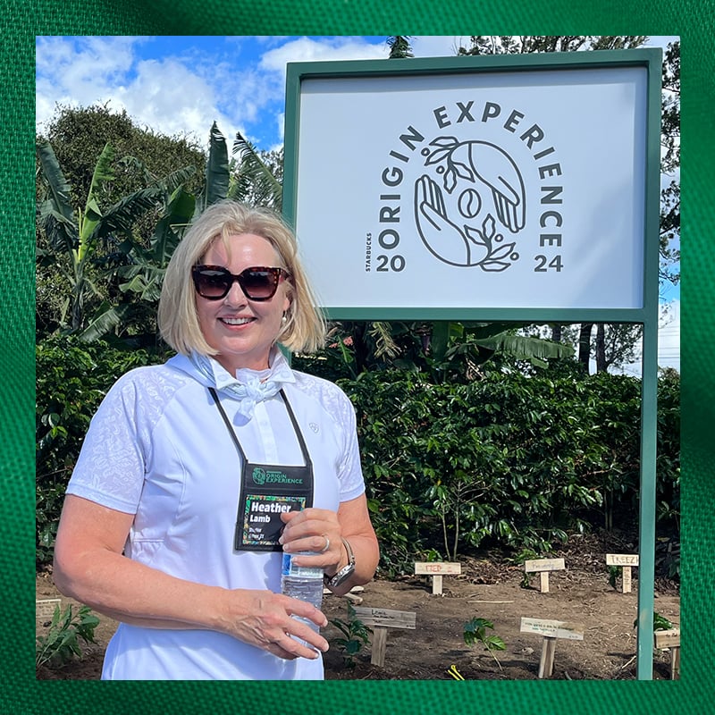 A woman with blonde hair, wearing sunglasses and a white shirt, stands in front of a sign that reads "Origin Experience 2024." She holds a water bottle and has a name tag around her neck. The background features lush greenery and small signs in the ground.