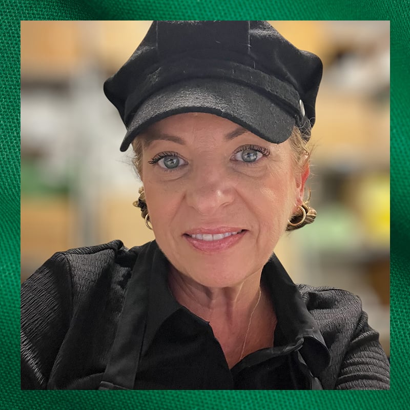 A woman with light skin and blue eyes is smiling, wearing a black cap and a black shirt. She has earrings and is in a kitchen setting.