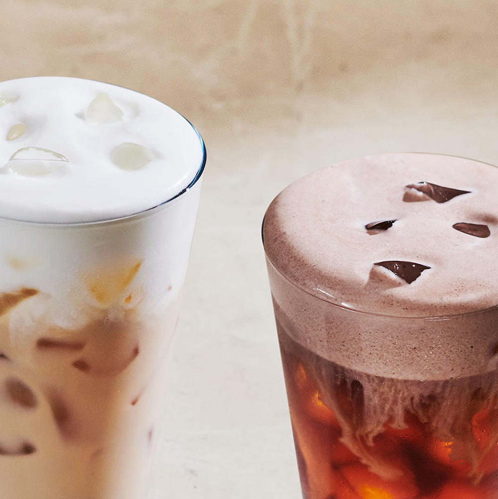 Two glasses of beverages are displayed. The left glass contains a creamy iced drink topped with foam and ice cubes, while the right glass features a dark iced drink with a frothy top and ice. The background is a soft, neutral tone.